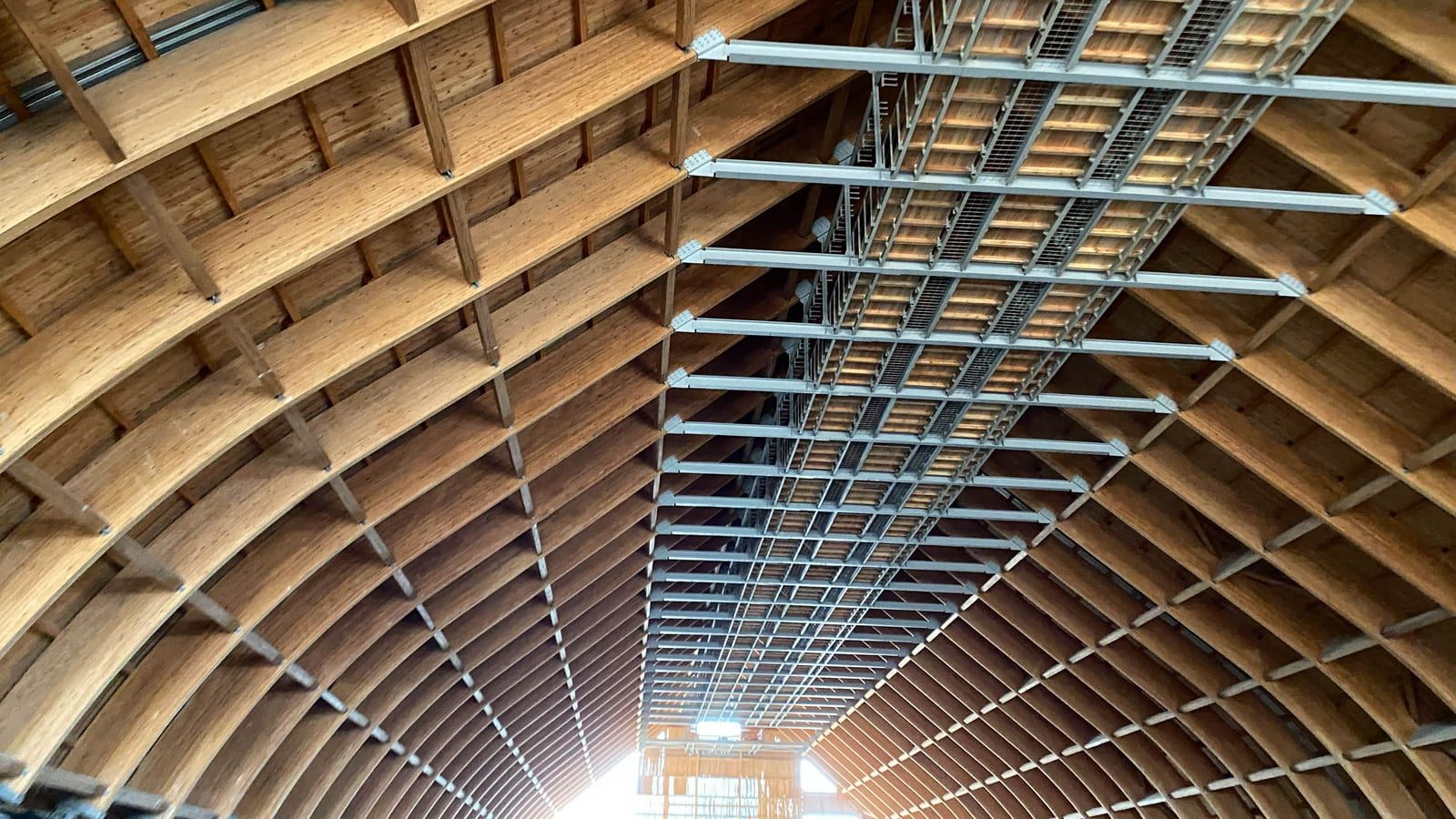Arcos de madera laminada de gran luz con correas de madera y una pasarela técnica metálica a lo largo de la cubierta, vistos desde abajo