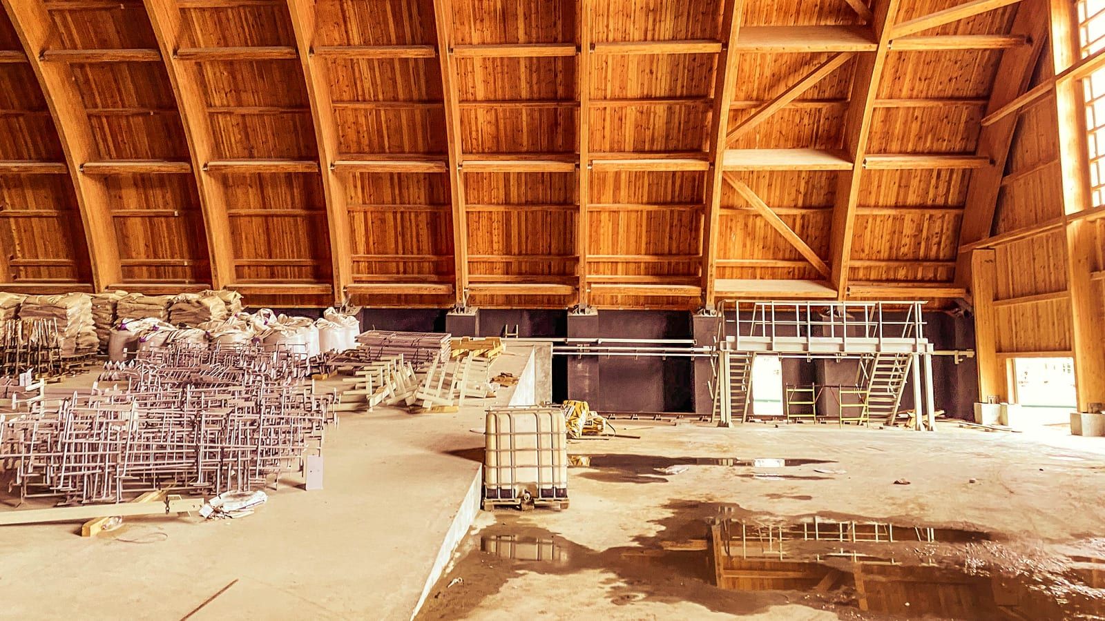 Nave en obra con arcos de madera laminada, cerramiento de tablas de madera y material de construcción en el suelo