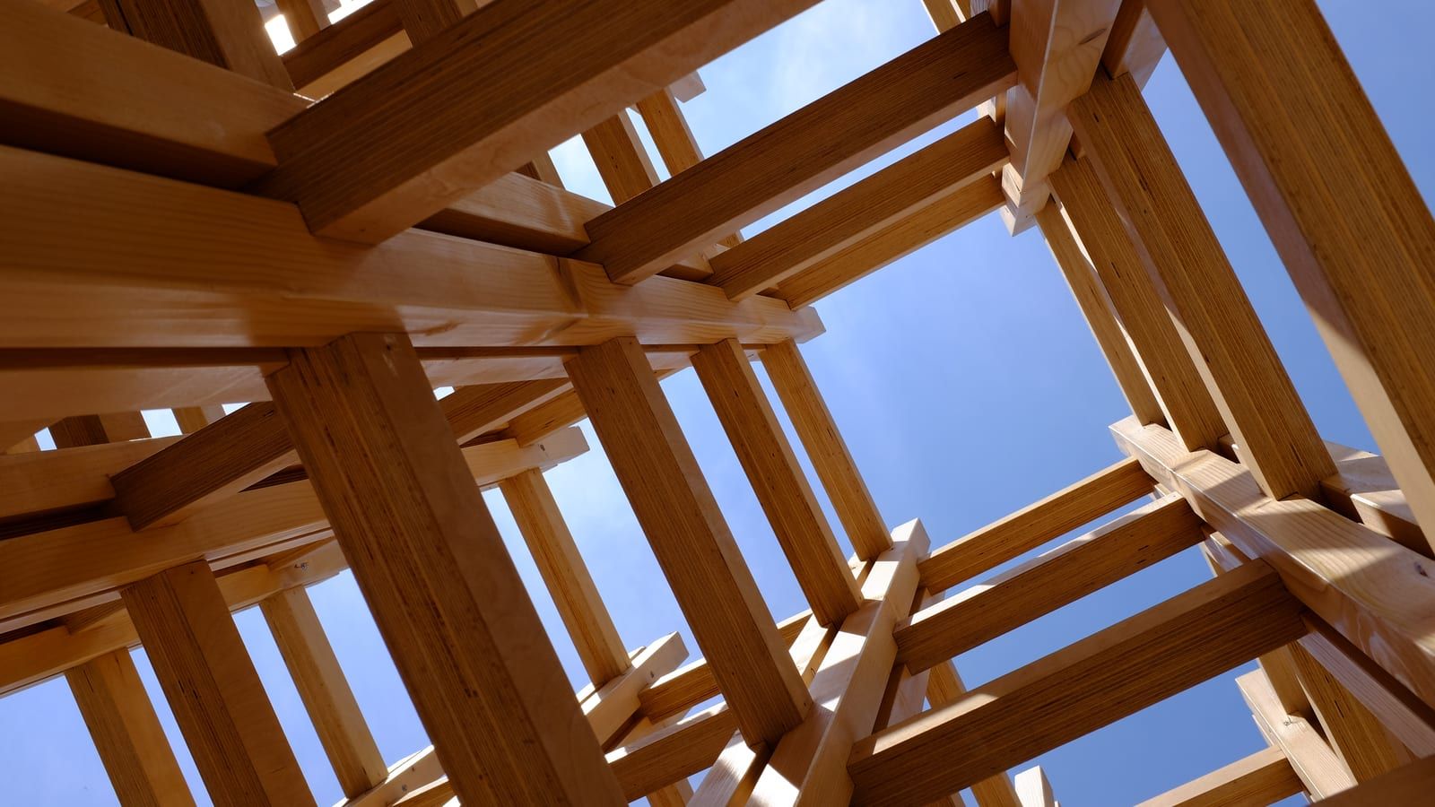 Estructura de vigas de madera entrecruzadas vista desde abajo, con el cielo azul al fondo