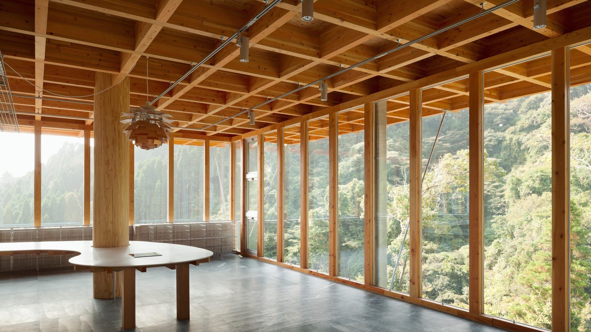 Sala de un edificio con retícula de vigas de madera en el techo, pilar de madera y fachada acristalada con montantes de madera